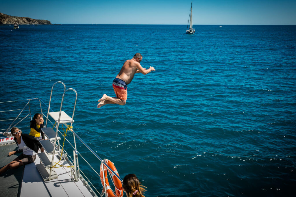 Un homme en plein saut depuis le plongeoir situé à l'avant du catamaran dans les eaux cristallines de la baie de Paulilles lors d'une Sortie bateau port-vendres