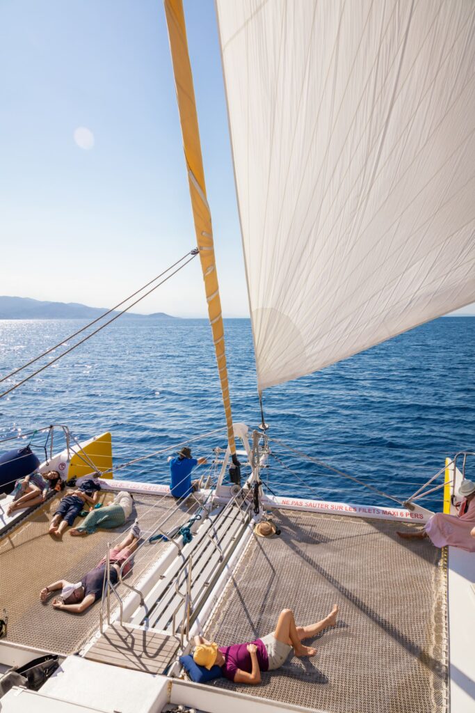 Des passagers se relaxant dans les filets avant du catamaran au retour d'une sortie pour savoir où voir des dauphins à Perpignan, naviguant au large de Cerbère et Banyuls-sur-Mer avec une vue panoramique sur le Cap Béar au fond, avec le catamaran Navivoile