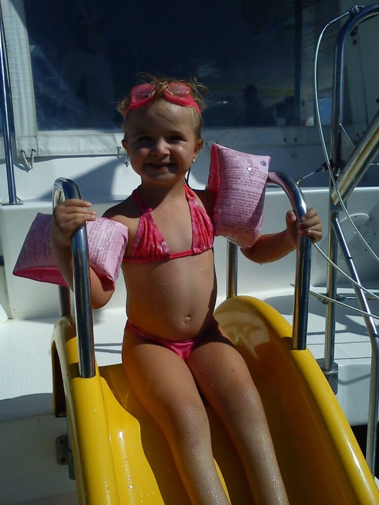 Une petite fille rayonnante avec des brassards aux bras, assise en haut du toboggan à l'arrière du catamaran Navivoile, s'apprêtant à glisser dans les eaux de la baie de Paulilles lors d'une Sortie bateau Port-vendres