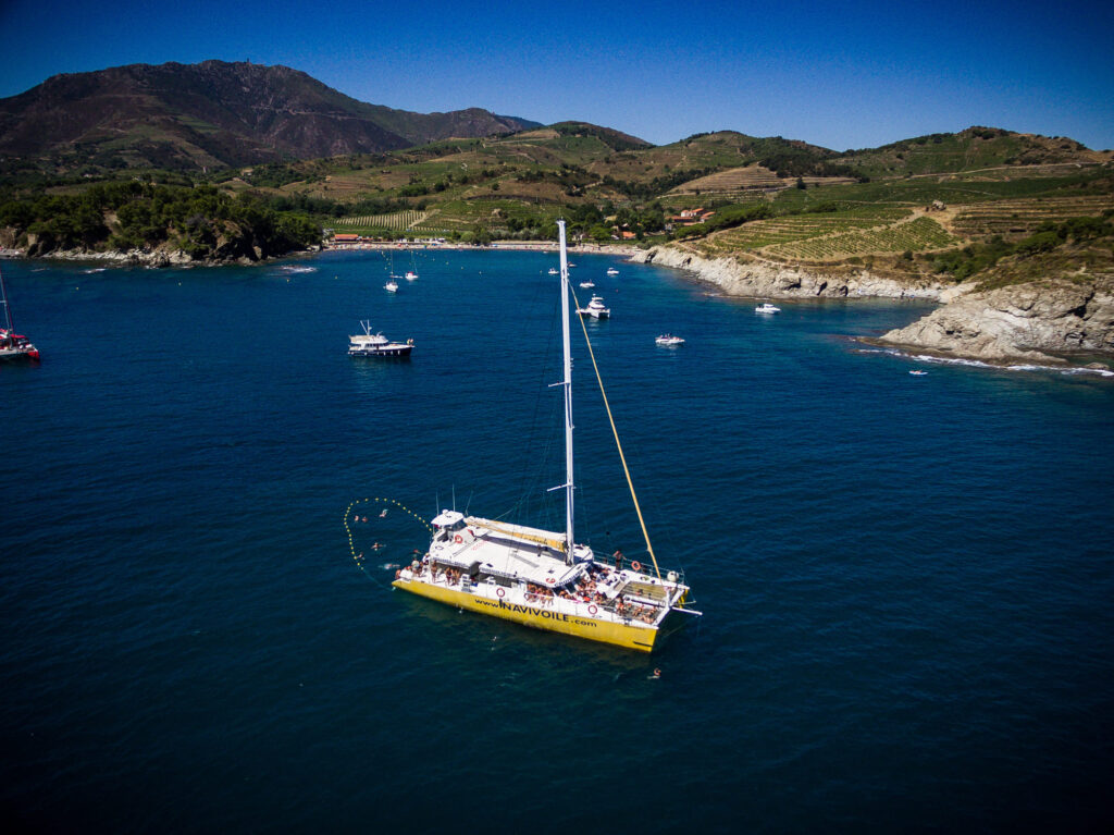 Le catamaran Navivoile à l'ancre dans les eaux turquoise de Paulilles, avec en arrière-plan le massif des Albères plongeant dans la mer, lors d'une sortie bateau à la journée à Canet