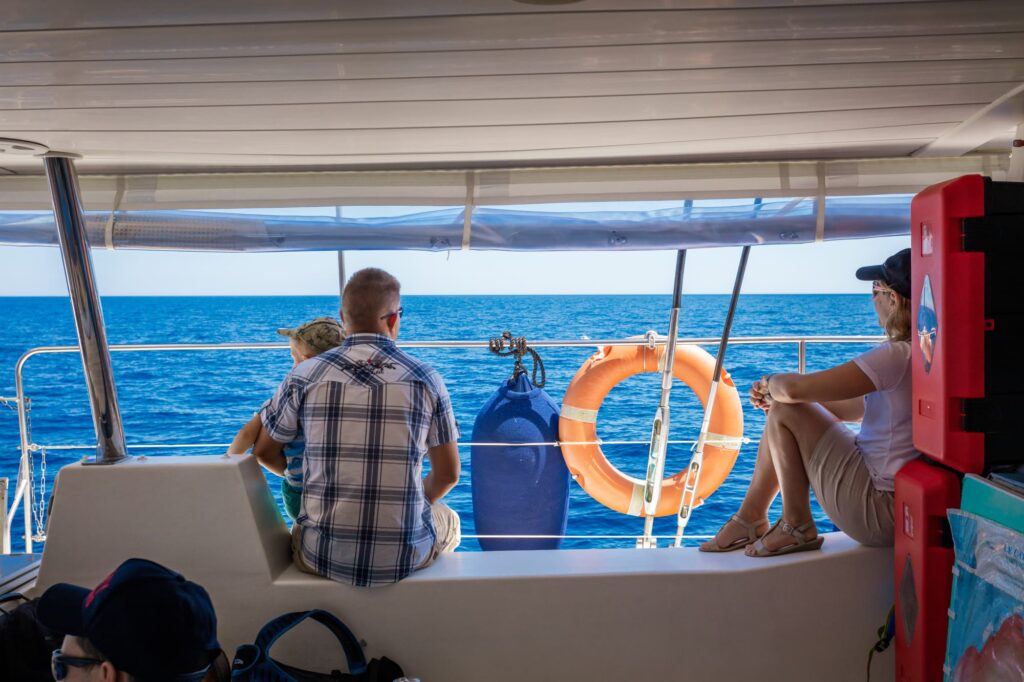 Vue de dos de deux adultes et un enfant observant sereinement l'horizon marin à l'abri du soleil sous la nacelle du catamaran lors d'une Sortie en mer à Canet-en-Roussillon