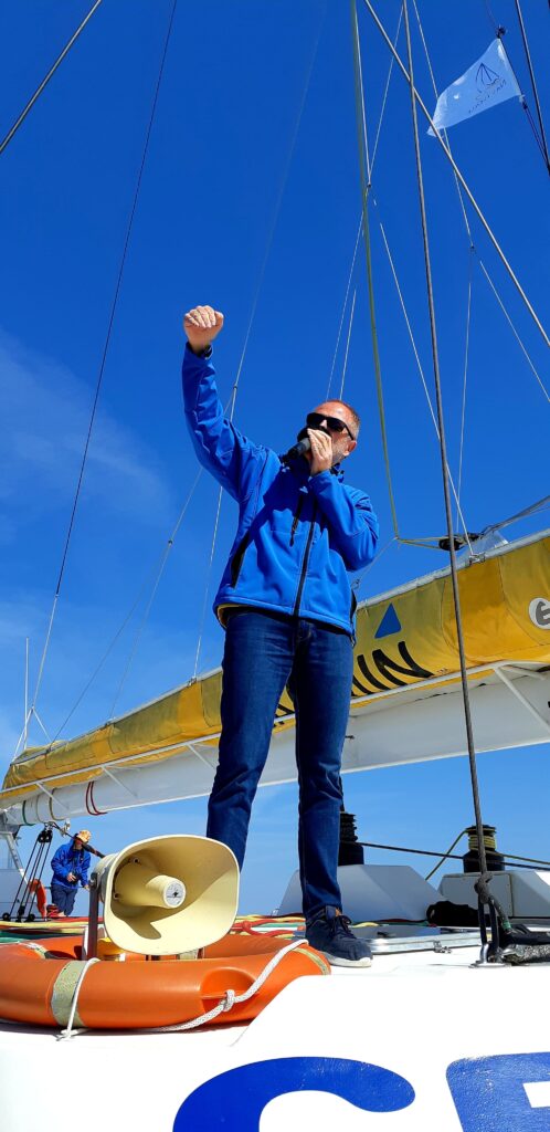 Photo du capitaine au poste de pilotage sur le roof lors d'une Promenade en mer à Canet en Roussillon. Il commente le panorama sur Perpignan et les Pyrénées sous voiles, face au littoral de Canet Sud, à bord du catamaran Navivoile